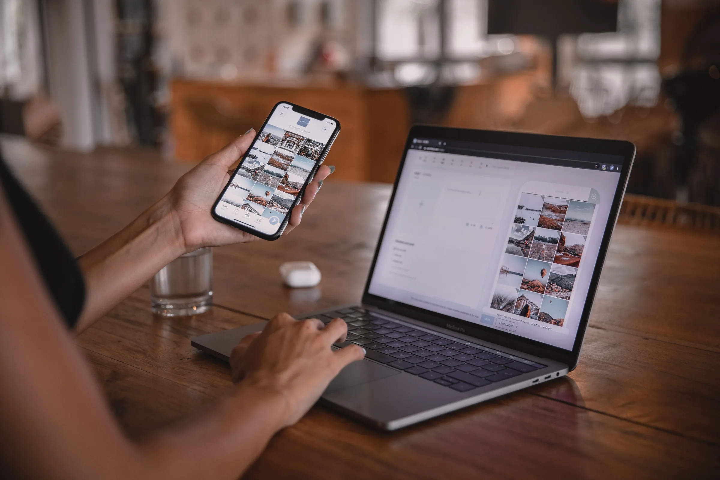 Person holding smartphone displaying photo gallery while working on laptop at wooden desk.