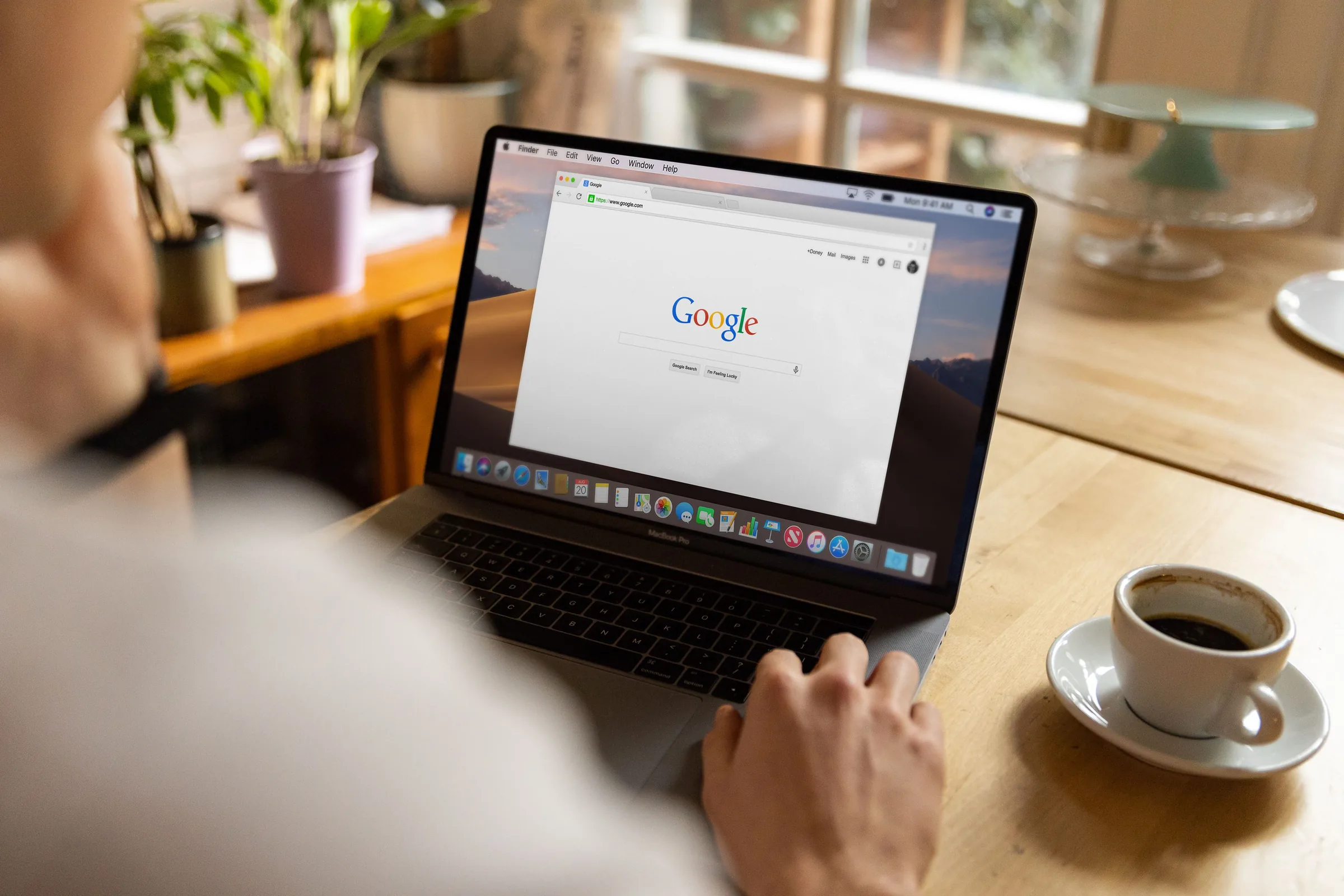 Person using laptop displaying Google search homepage at wooden desk with coffee cup and potted plants.