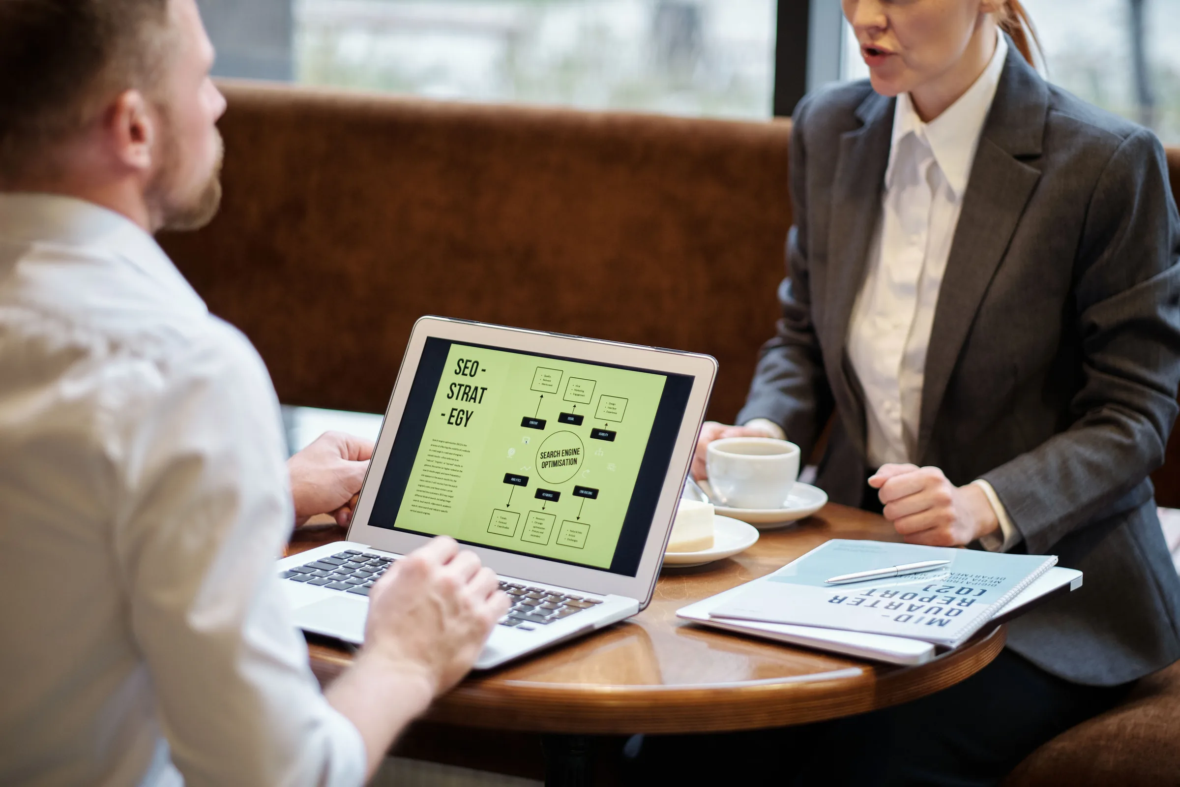 Two business professionals discuss SEO strategy on a laptop during a meeting at a wooden table.