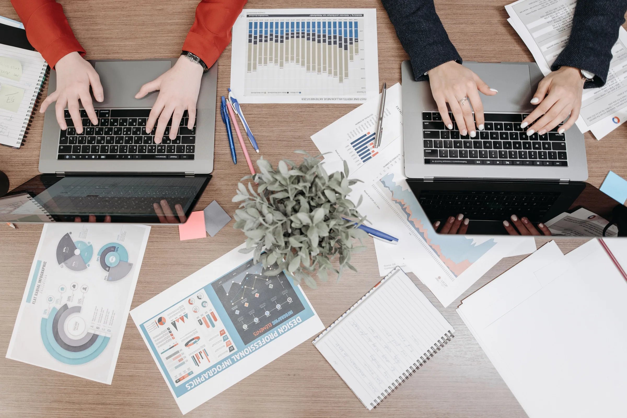 Two marketing professionals working on laptops surrounded by data charts and analytics reports on a wooden desk.